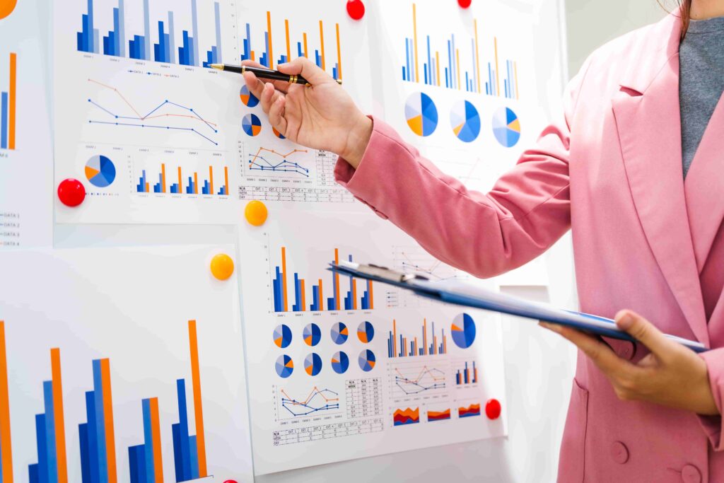 A businesswoman in a pink suit points at a board filled with colorful bar charts and pie graphs, illustrating data analysis for improved insurance customer experience and business growth.