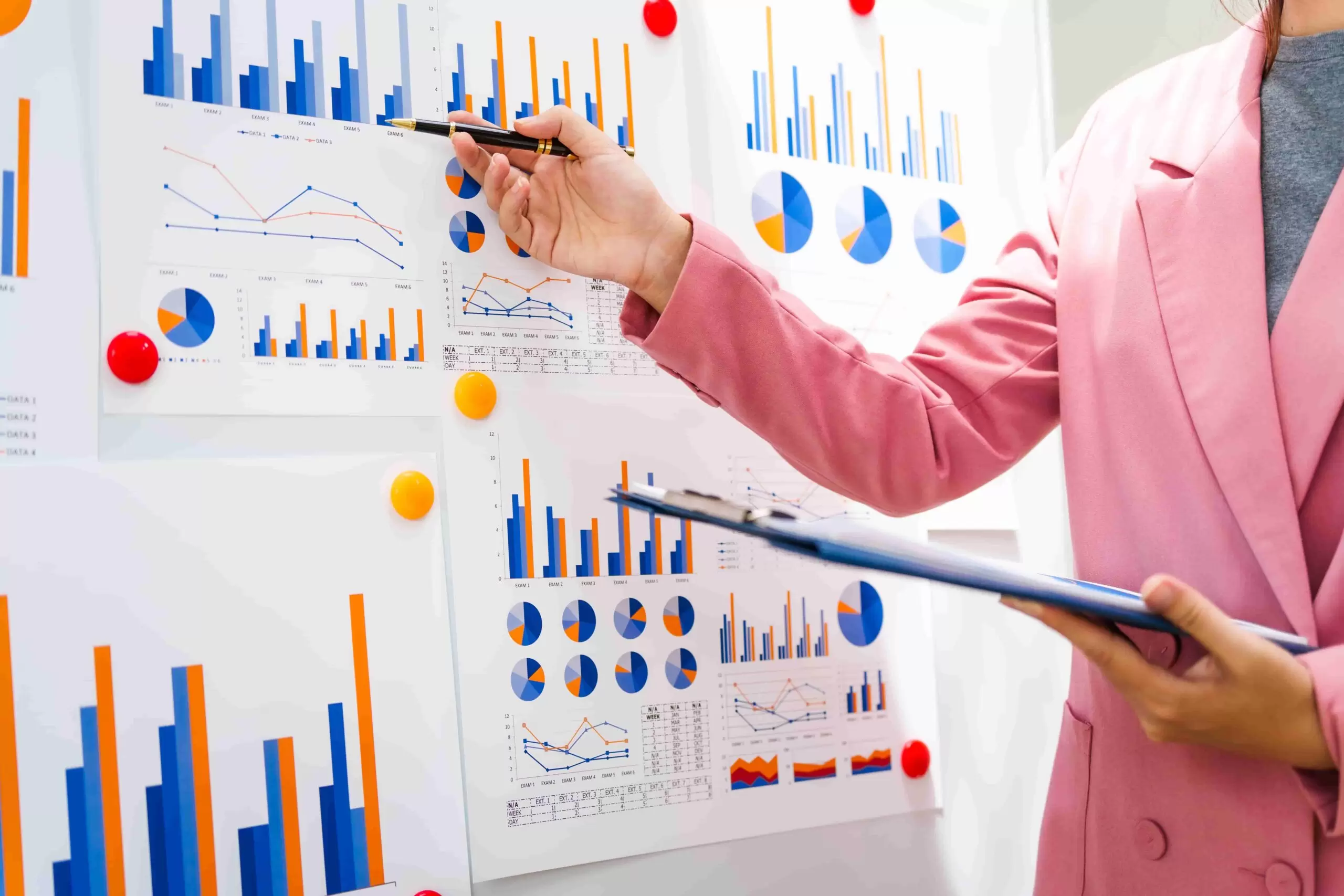 A businesswoman in a pink suit points at a board filled with colorful bar charts and pie graphs, illustrating data analysis for improved insurance customer experience and business growth.