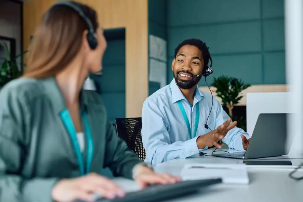 A young Black businessman wearing a headset and a blue shirt is seated at a desk, smiling and gesturing with his hand while looking at a female colleague who is out of focus in the foreground. He has a laptop open in front of him. The image captures a moment of friendly and efficient communication, showcasing a diverse team member providing attentive customer service in insurance.