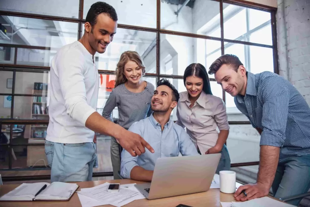 A team of diverse and smiling professionals gathered around a laptop, collaborating and pointing at the screen, representing efficient teamwork and expertise in outsource underwriting services.