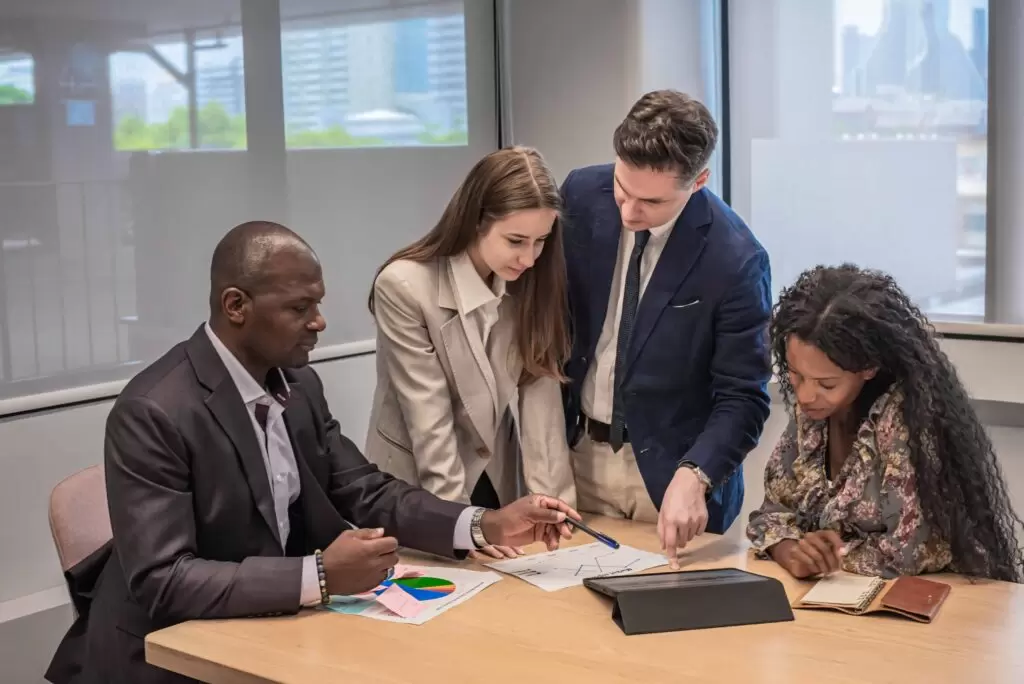 A diverse group of business professionals meets around a table, using both a tablet and paper documents to discuss and review an end-to-end claims processing workflow.