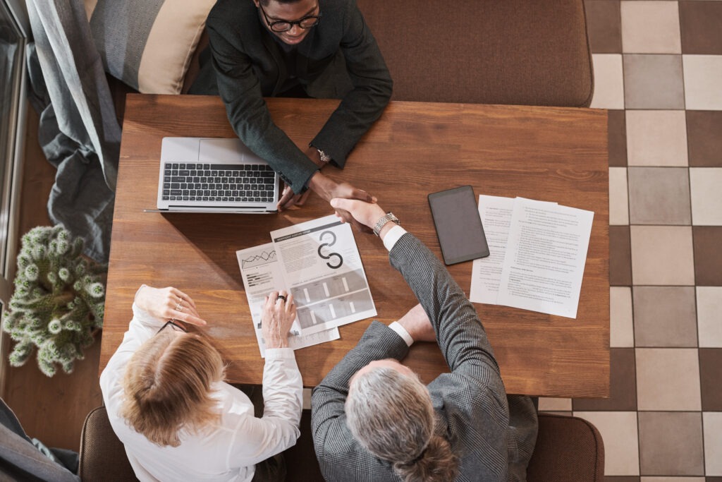 Two business people shake hands over a table with a laptop and documents, symbolizing the final stages and successful conclusion of an end-to-end claims process.
